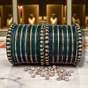 Puede incluir: Un conjunto de brazaletes verde oscuro con detalles dorados y adornos brillantes. Una dispersión de pequeñas piedras preciosas transparentes se encuentra frente a los brazaletes sobre una superficie de mármol blanco. Los brazaletes probablemente son parte de una colección de joyas.