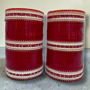 May include: Two stacks of red bangles with white and gold decorative bands. The bands are adorned with small, sparkling rhinestones. The bangles are a traditional accessory, often worn for special occasions.