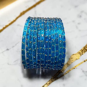 May include: A collection of blue bangles with gold detailing. The bangles are a bright blue colour and seem to be made of a transparent material. The gold accents are small and evenly spaced along the bangles.