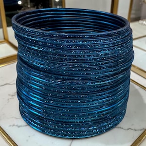 May include: A collection of teal blue bangles with a shimmering, glittery surface. The bangles are stacked together, creating a cylindrical shape. The jewellery is displayed on a white and grey surface.