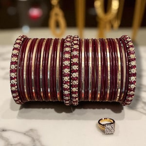 Puede incluir: Una pila de brazaletes granate y dorados con detalles de pedrería. Un anillo de oro con una piedra cuadrada con forma de diamante está delante de los brazaletes. Las joyas se exhiben sobre una superficie clara.