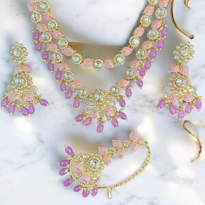 Puede incluir: Un conjunto de joyas dorado que incluye un collar, pendientes y una pieza para la cabeza. Las piezas están adornadas con detalles de esmalte rosa en forma de hoja, piedras preciosas transparentes y cuentas moradas colgantes. El conjunto se presenta sobre una superficie de mármol blanco.