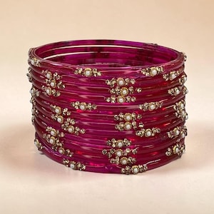 May include: A stack of translucent magenta bangles featuring small, circular white beads and gold-toned metallic accents arranged in floral-like patterns along the outer surface of each individual bracelet in the set.