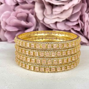 Könnte beinhalten: Satz von sechs goldenen Armreifen mit klaren Strasssteinen und rosa quadratischen Steinen.