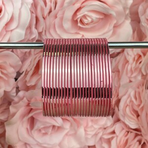 Op de afbeelding: Een stapel roze en gouden bangles met een glanzende metallic afwerking. De bangles zijn verticaal gestapeld en worden getoond tegen een achtergrond van roze rozen.