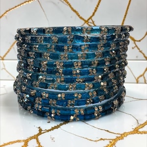 May include: A stack of ten circular bangles in a deep teal blue colour. Each bangle features a repeating pattern of rectangular blue segments interspersed with small, clear, sparkling crystals arranged in clusters around the entire circumference.