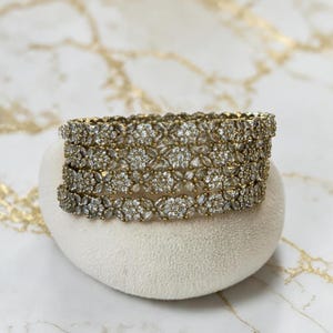 May include: A set of gold-coloured bangles featuring a floral pattern, adorned with clear, sparkling gemstones. The bangles are presented on a white, heart-shaped surface against a marble backdrop.