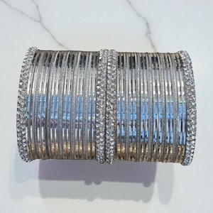 May include: A stack of silver bangles with a sparkling rhinestone border. The bangles are arranged in a neat stack, reflecting light and creating a shimmering effect. The jewelry is displayed on a white marble surface.