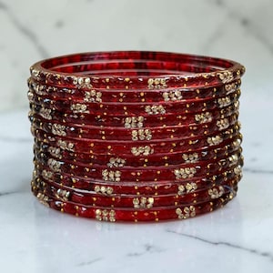 May include: A stack of deep red bangles featuring a faceted texture and embedded with small, clear, sparkling crystals arranged in repeating patterns along the circumference of each individual circular bracelet.