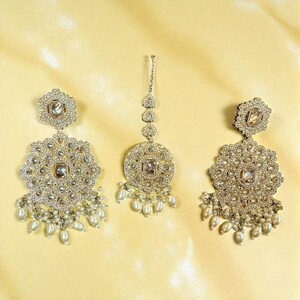 May include: A silver-toned jewellery set comprising earrings and a headpiece. The earrings and headpiece are embellished with clear crystals and faux pearls. The earrings have a large, circular design with dangling pearls. The headpiece features a central pendant and a chain.