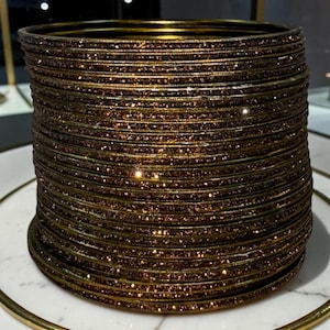 May include: A collection of brown and gold bangles with a glittery finish. The bangles are stacked on a white and gold decorative plate. The jewellery has a metallic sheen and a classic design.