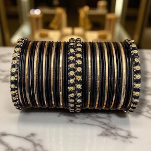 May include: A stack of black and gold bangles. The bangles are a mix of solid black and gold-toned metal, with decorative bands of gold and small, sparkling embellishments. The stack is displayed on a marble surface.