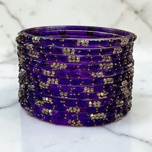 May include: A stack of deep purple translucent bangles featuring a faceted texture and embedded clear crystal accents. Each circular bracelet is uniform in size and design, creating a cohesive set of jewellery with a reflective, polished finish.