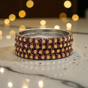 May include: Four silver bangles, each featuring alternating rows of small, round, ruby-red and yellow gemstones. The bangles are stacked on a reflective surface, with blurred bokeh lights in the background.