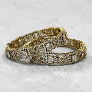 May include: Two gold-toned bangles featuring a repeating pattern of rectangular-cut clear stones interspersed with clusters of small, round, sparkling white stones. The metal surface has a textured, intricate design surrounding each stone setting.