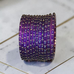 May include: A collection of purple bangles with a faceted design and gold-coloured accents. The bangles are stacked together, displaying their intricate details and rich colour. The background is a light, neutral surface.
