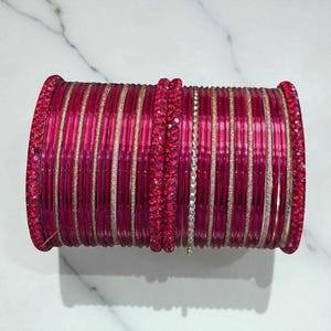 Op de afbeelding: Een stapel felroze armbanden met gouden accenten. De armbanden zijn versierd met sprankelende rode versieringen op de randen en een enkele zilveren armband met heldere stenen. De sieraden worden gepresenteerd op een wit marmeren oppervlak.