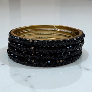 May include: A set of black beaded bangles with a gold-coloured inner band. The bangles are stacked together, displaying the intricate details of the black beads. The gold band adds a touch of elegance to the jewellery.