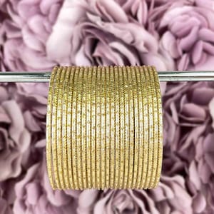 Peut inclure: Une pile de bracelets dorés avec une finition scintillante. Les bracelets sont disposés en cercle et sont présentés sur un fond de fleurs violettes.