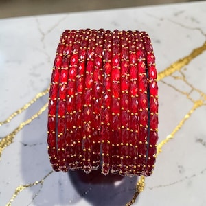 May include: A collection of red beaded bracelets. Each bracelet is made of small, faceted red beads with gold-coloured accents. The beads are arranged in rows, creating a textured surface. The bracelets are displayed on a white and gold surface.