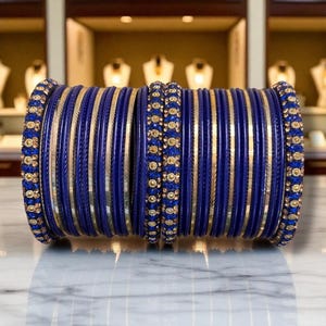 May include: A stack of blue and gold bangles. The bangles are a deep blue color with gold accents. Some bangles have gold and crystal embellishments. The bangles are stacked on a white surface.