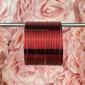 May include: A collection of red bangles, set against a backdrop of pink roses. The bangles are slender and seem to be made of glass or a similar material, reflecting light. The overall aesthetic is delicate and feminine.
