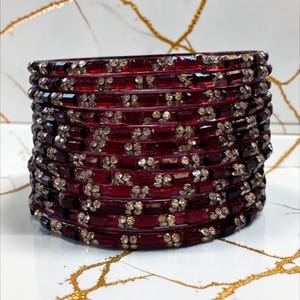 May include: A stack of deep red bangles featuring a repeating pattern of small, clear, sparkling stones embedded along the surface of each individual bracelet. The bangles are uniform in colour and design, creating a cohesive, layered appearance.