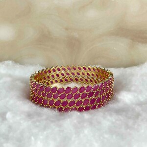 May include: A gold-coloured bangle bracelet featuring a repeating pattern of oval, deep pink gemstones. The bracelet is set against a soft, white background, highlighting the jewellery's design and colour.