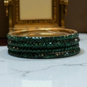 May include: Four green bangles with a gold band, stacked together. The bangles are adorned with small, sparkling green stones. A gold-framed mirror is in the background.