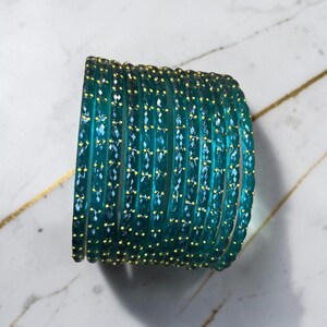 May include: A collection of teal glass bangles with a faceted design and small gold-coloured accents. The bangles are arranged on a white and gold marble surface. The jewellery is a fashion accessory.