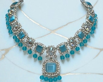 May include: A silver-toned necklace with a central square pendant and matching square and round blue gemstones. The necklace features a series of smaller blue beads and clear crystals, creating a sparkling, elegant design. The necklace is displayed on a light blue surface.