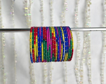 May include: A collection of thin, circular bangles arranged in a row on a horizontal metal rod. Each bangle features a vibrant, multi-colored tie-dye pattern in shades of blue, yellow, green, red, and pink, accented with small, evenly spaced gold dots.