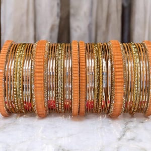 May include: A display of gold and orange bangles. The bangles are arranged in a cylindrical form, with alternating gold and orange bands. Some bangles feature small, sparkling decorations. The background is a marble surface with a gold frame.