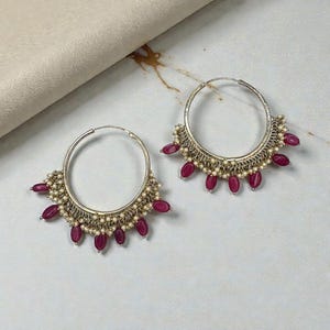 Puede incluir: Un par de pendientes de aro plateados con un borde inferior decorativo. Los pendientes presentan pequeñas cuentas blancas y piedras en forma de lágrima de color granate. Los pendientes se exhiben sobre una superficie clara.