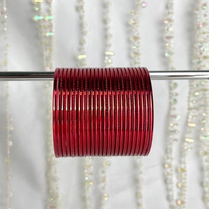 May include: A collection of red bangles, presented on a silver bar. The bangles have a rich, glossy red hue, reflecting light. The background shows strands of white beads, providing a soft, diffused backdrop. These are traditional Indian jewellery.