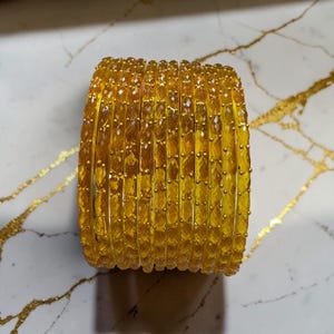 Op de afbeelding: Een stapel gele armbanden met een facetontwerp. De armbanden hebben een warme, gouden kleur en lijken gemaakt te zijn van glas of een vergelijkbaar materiaal. De armbanden zijn op elkaar gestapeld, waardoor een solide, cilindrische vorm ontstaat. De achtergrond is een marmeren oppervlak.