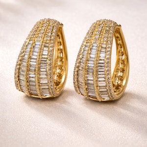 May include: A pair of gold-coloured hoop earrings. Each earring is encrusted with rows of baguette-cut and round-cut clear gemstones. The earrings have a polished finish and are displayed on a light-coloured surface.