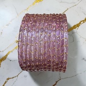 May include: A collection of light purple bangles with a faceted design. Each bangle features small, evenly spaced gold-coloured beads. The bangles are stacked together, creating a layered effect. The background is a marble surface.