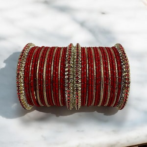 Könnte beinhalten: Ein Stapel roter Armreifen mit goldenen und Strass-Akzenten. Die Armreifen sind zylindrisch angeordnet, mit einer Mischung aus roten und goldgestreiften Bändern. Die Enden und die Mitte weisen aufwendige Strassdetails auf, die ein festliches und verziertes Aussehen erzeugen.