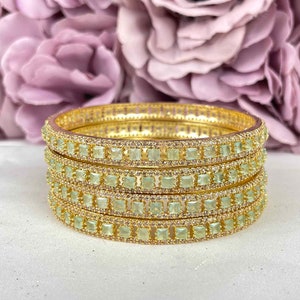 Op de afbeelding: Een stapel van vier goudkleurige armbanden. Elke armband is versierd met rijen kleine, vierkante, lichtgroene edelstenen en heldere kristallen. Zachte, wazige paarse bloemen op de achtergrond.