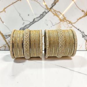 Może przedstawiać: Dwa zestawy złotych bransoletek typu bangle, z których każda zawiera kombinację gładkich metalowych obręczy, teksturowanych obręczy pokrytych brokatem oraz cienkich obręczy owiniętych drutem. Bransoletki są ułożone w dwa oddzielne stosy.