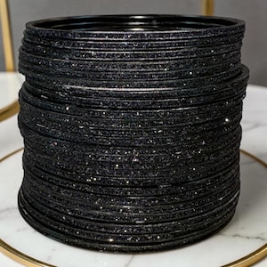 May include: A stack of black bangles with a glittery, textured surface. The bangles are stacked on a white and gold decorative plate. The bangles are a dark, solid color.