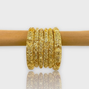 Könnte beinhalten: Sieben goldene Armreifen mit aufwendigen floralen und geometrischen Mustern werden auf einem Holzstab präsentiert. Die Armreifen sind stark reflektierend, und ihre detaillierten Designs sind sichtbar. Der Hintergrund ist eine schlichte weiße Oberfläche.