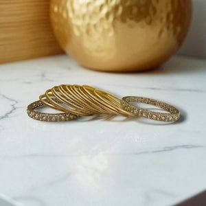 May include: A collection of gold bangles, some adorned with small, sparkling embellishments. The bangles are arranged on a white marble surface, with a large gold decorative object in the background. The overall aesthetic is elegant and sophisticated.
