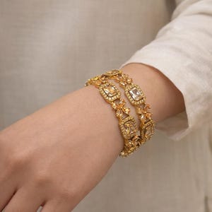 Könnte beinhalten: Zwei goldfarbene Armreifen mit quadratischen und floralen Akzenten. Die Armreifen sind mit klaren, quadratisch geschliffenen Edelsteinen besetzt. Der Schmuck wird am Handgelenk vor einem neutralen Hintergrund getragen.