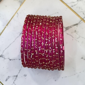 May include: A collection of ruby-red bangles with a faceted design and gold-coloured accents. The bangles are arranged in a circular shape, highlighting their intricate details and reflective surfaces. The jewellery is set against a marble background.