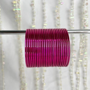 Op de afbeelding: Een stapel levendige magenta armbanden, gepresenteerd op een zilveren staaf tegen een zachte, vage achtergrond. De armbanden zijn dicht op elkaar gepakt en vertonen een glanzende afwerking. De afbeelding benadrukt de kleur en textuur van de sieraden.