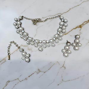 May include: A silver-toned jewelry set featuring a necklace, earrings, and a pendant. The necklace and earrings have a cluster design with faux pearls and clear crystals. The pendant has a similar design and is attached to a chain.