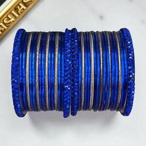 Puede incluir: Una pila de brazaletes azules con una apariencia brillante y similar a una joya. Los brazaletes son de un color azul vibrante, con una banda plateada entre cada brazalete. Los bordes de los brazaletes también están adornados con joyas azules.
