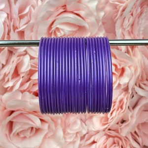 Puede incluir: Un conjunto de brazaletes de metal morado apilados uno encima del otro. Los brazaletes son brillantes y tienen un acabado liso.